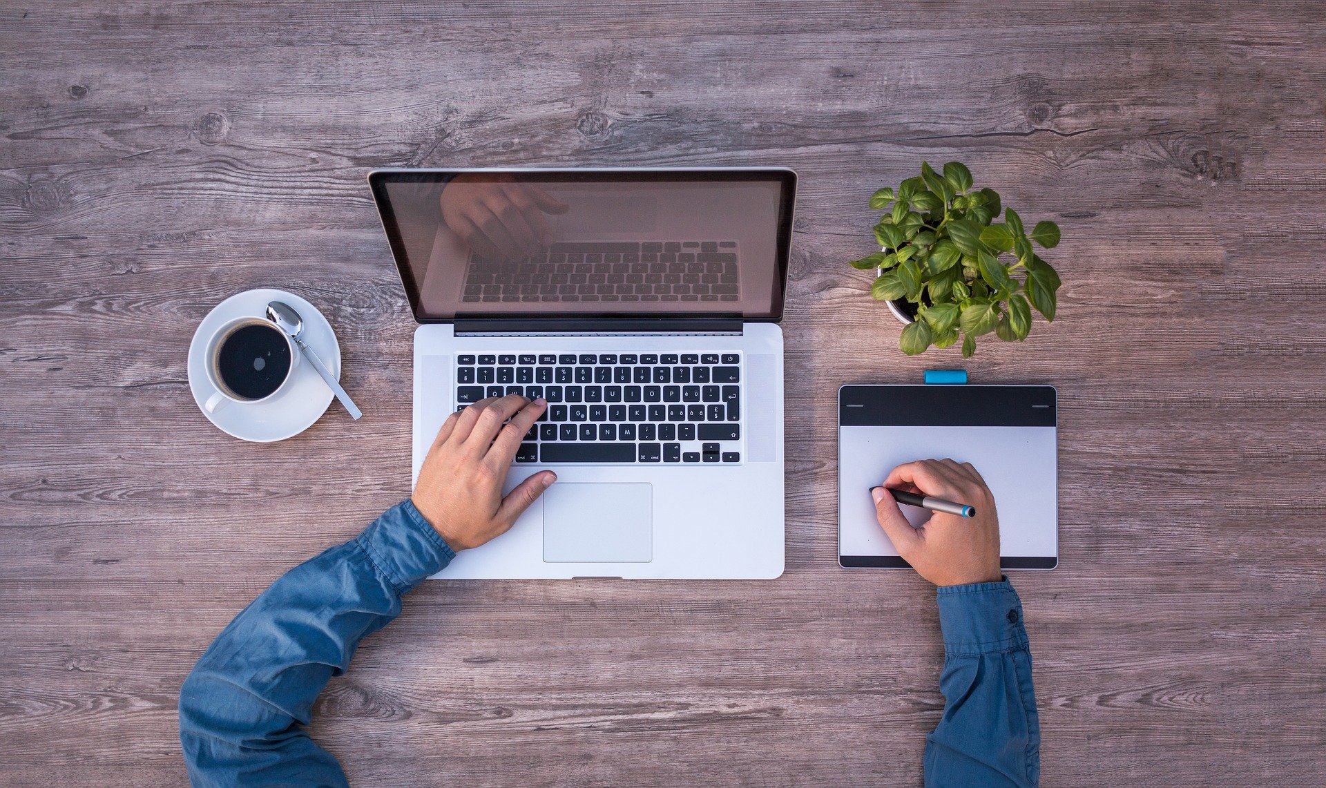 Top view of a gray wooden table and the hands of a working man. In front of him, an open laptop, a tablet, a cup of coffee and a flowerpot with a green plant